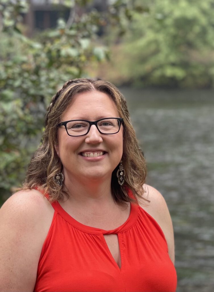 An image of Stephanie Schaefer, Psy.D. wearing glasses, an orange dress, looking out at the viewer with water and trees in the background. | Licensed psychologist Stephanie Schaefer in Portland, Oregon